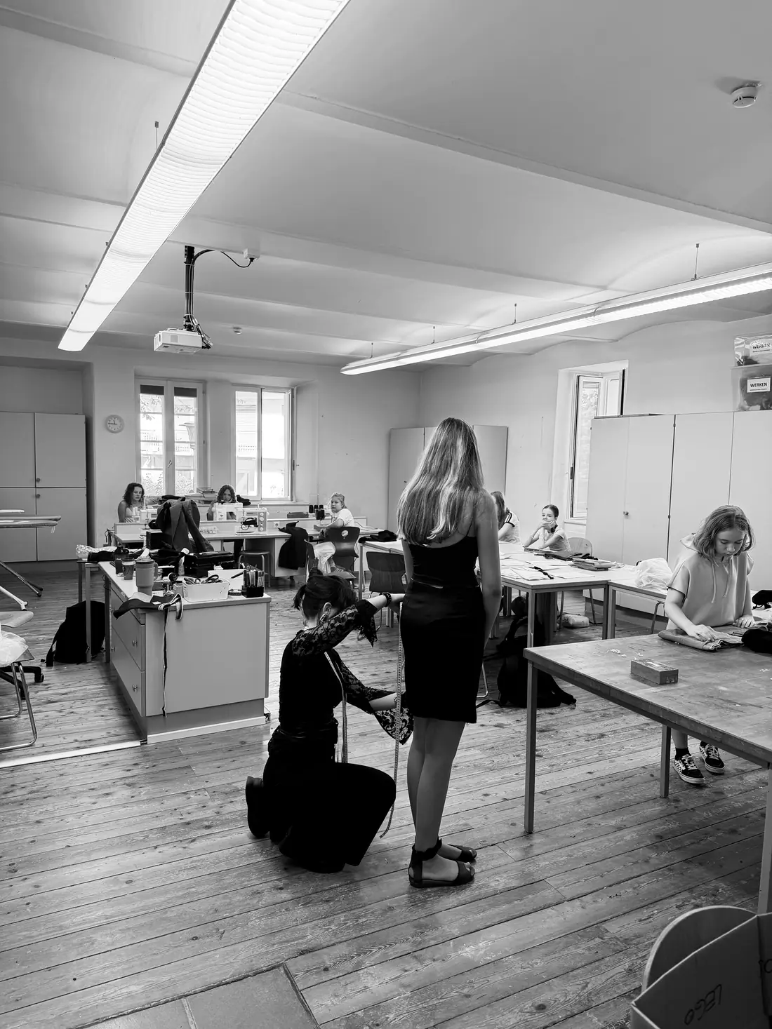 A fitting in the workshop room: a participant pins the hem of a dress while a classmate stands still.